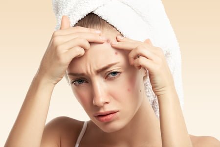 Headshot of displeased young blond woman with towel on her head, looking with painful face at the camera while squeezing pimple on her forehead. Portrait of Caucasian girl against blue wall background