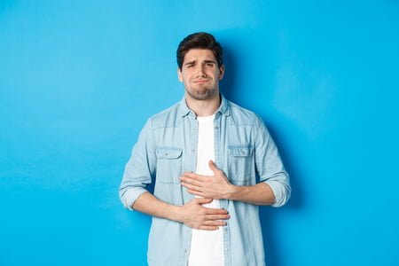 Man holding hands on belly and grimacing from pain, complaining on stomach ache, standing against blue background.