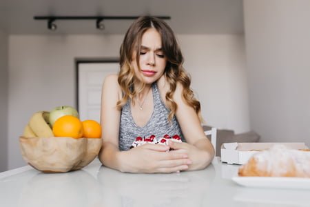 Sad young woman with curly hairstyle looking at cake. Indoor photo of pensive girl expressing negative emotions during diet.