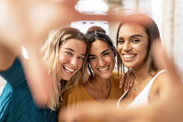 Three multiracial female friends taking selfie and making hand frame gesture in the street, looking through fingers at camera - Diverse young women having fun on vacation - Millennial people concept