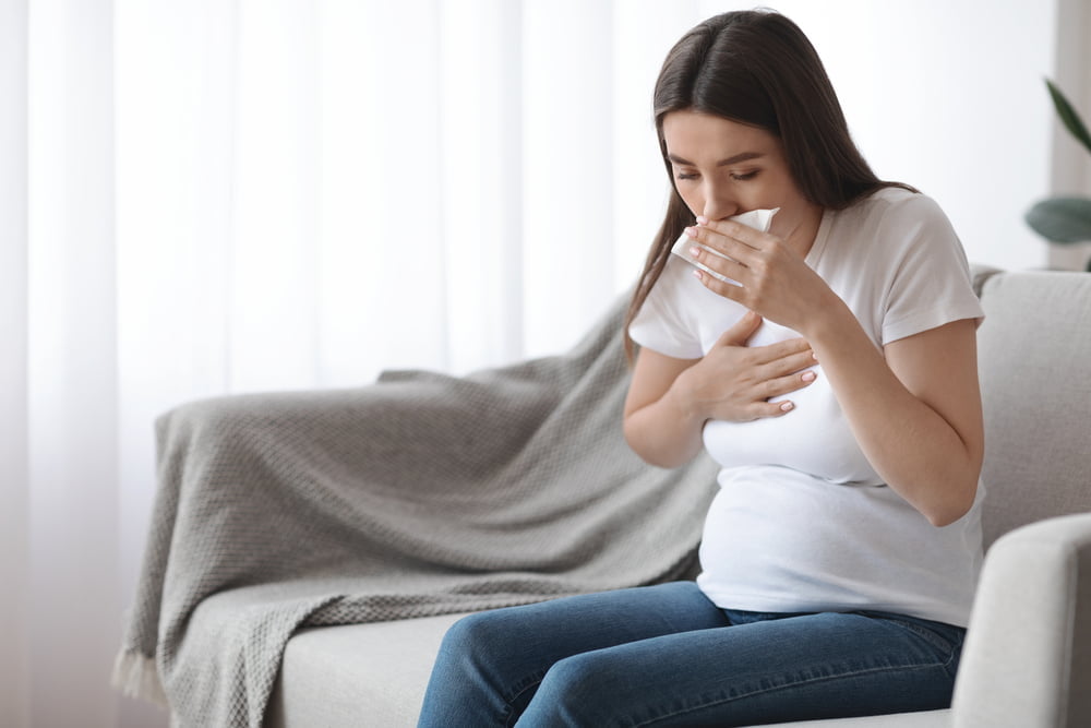 Pregnancy And Illness. Sick pregnant woman sitting on couch and coughing into tissue. Early pregnancy symptoms