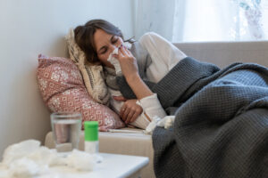 Sick woman lying on couch under the coverlet, wrapped in scarf, having flu or cold symptoms, sneezing in napkin. Flu.