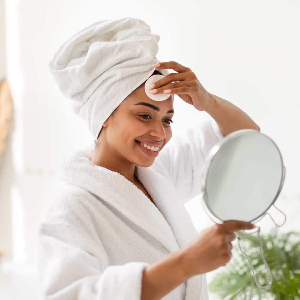 Black Woman Posing with Cotton Pad and Mirror Doing Facial Skincare Routine Removing Makeup Standing in Bathroom at Home. Female Caring for Face Skin Indoors. Beauty Care Concept. Selective Focus. Skin care routine. Managing oily skin.