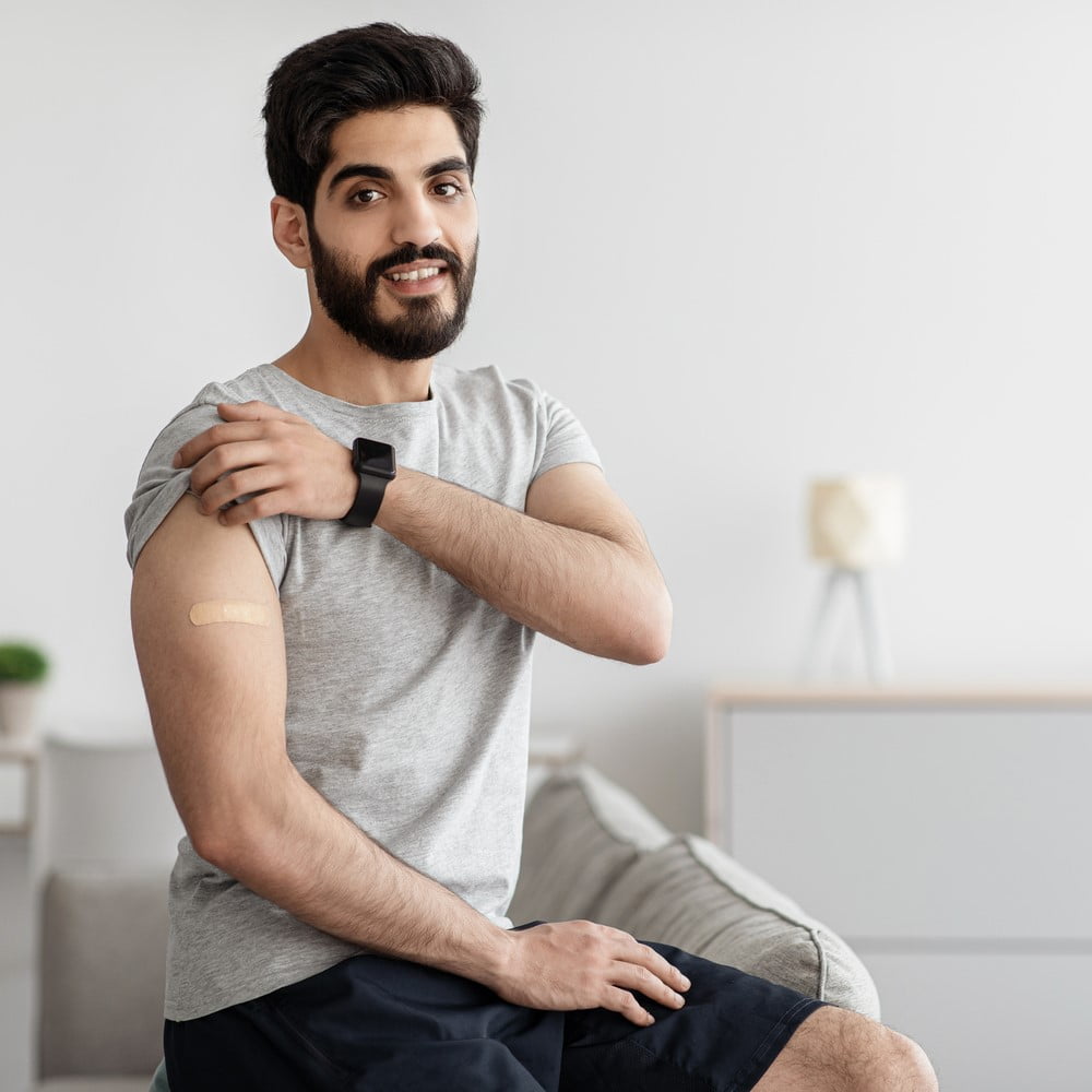 Recommendations for vaccination and immunization of population, health care concept. Cheerful Arabian bearded young male shows hand with band aid after injection in living room interior, empty space. Why preventive care is important concept. Immunization.