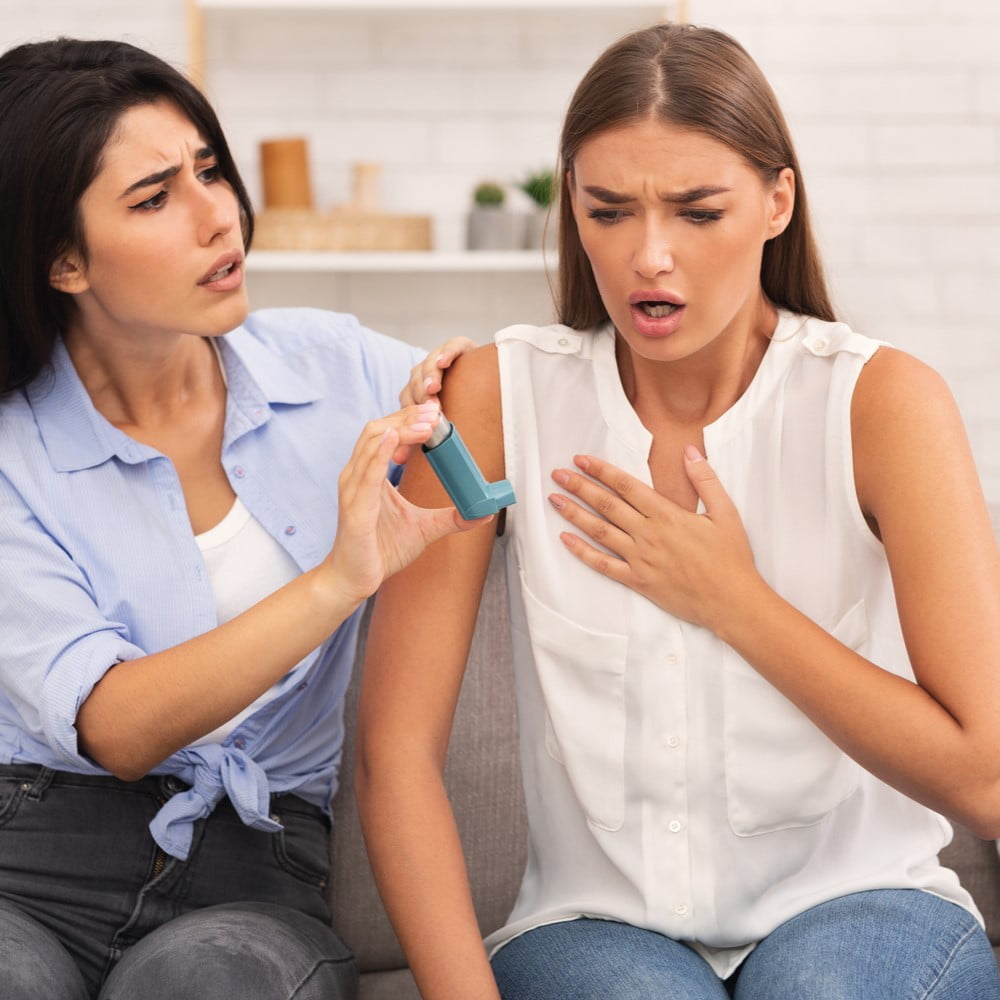 Girl Giving Asthma Inhaler to Friend Having Asthmatic Atack or Respiratory Depression Sitting On Couch At Home. Selective Focus. How asthma feels like, what asthma looks like.