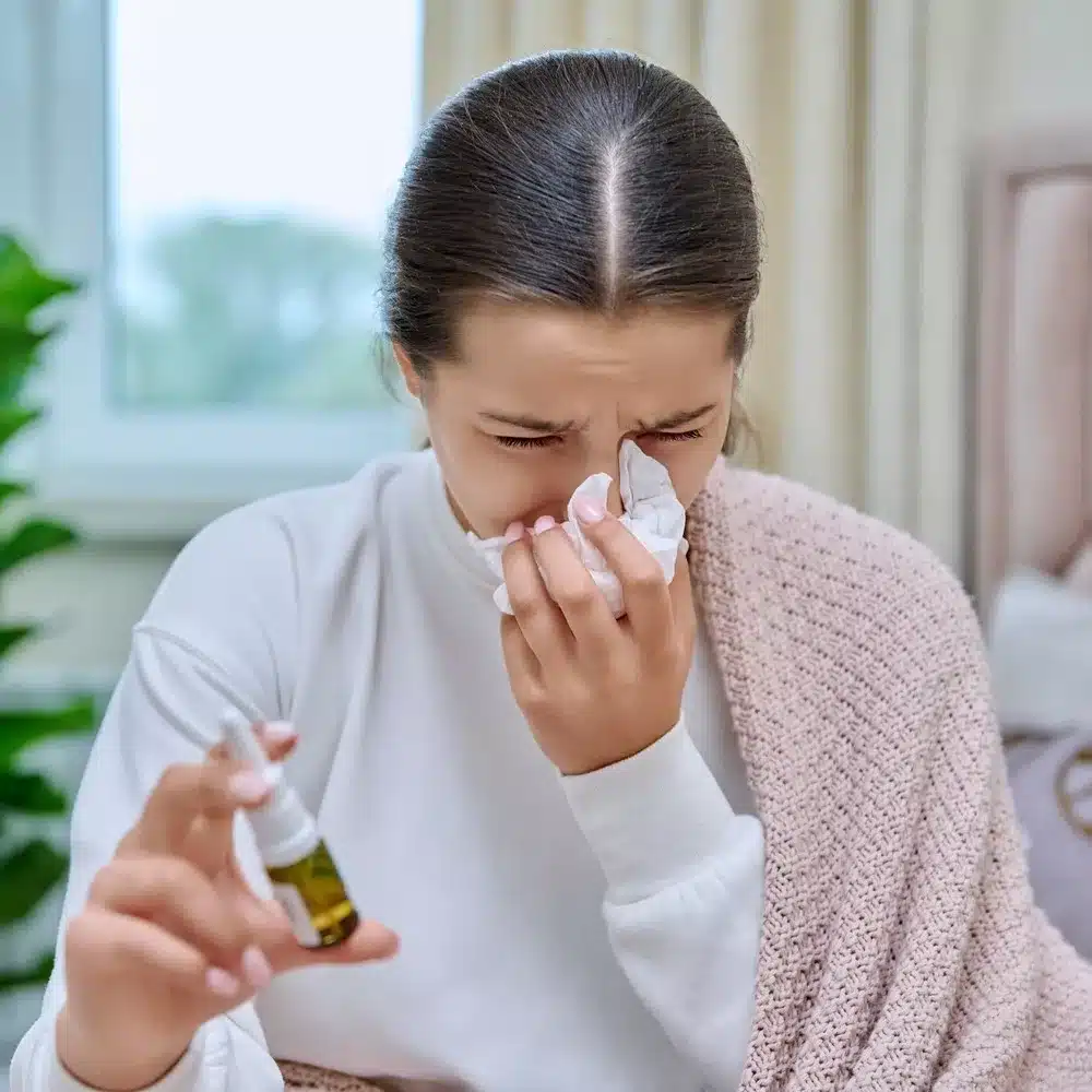 Flu season, autumn winter cold. Young teenage girl with runny nose treating rhinitis using nose drops spray. Allergic rhinitis symptoms. Sneezing.