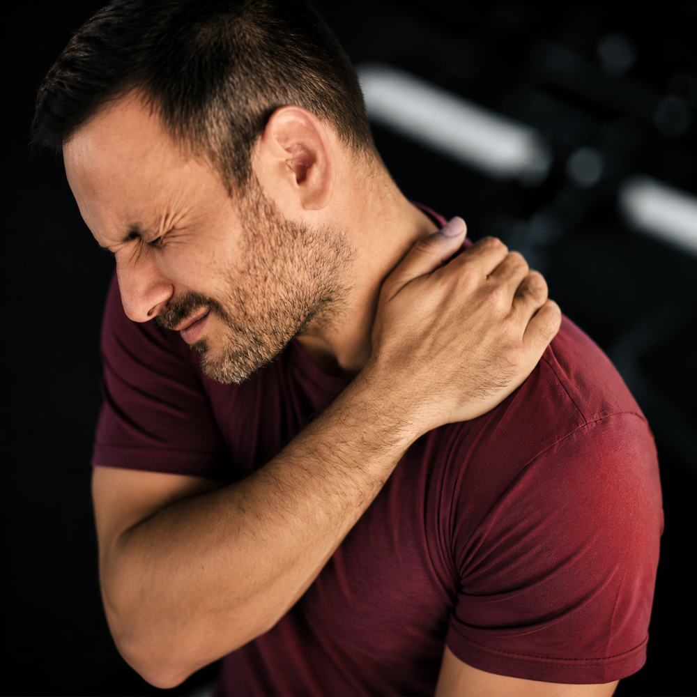 A man in a plum shirt holds his right shoulder and neck, with his eyes closed in discomfort. His dark hair is short, and he has a beard. This medium shot has a dark blurred background. This photo could be used for projects about pain management, exercise recovery, or physical therapy. Pain